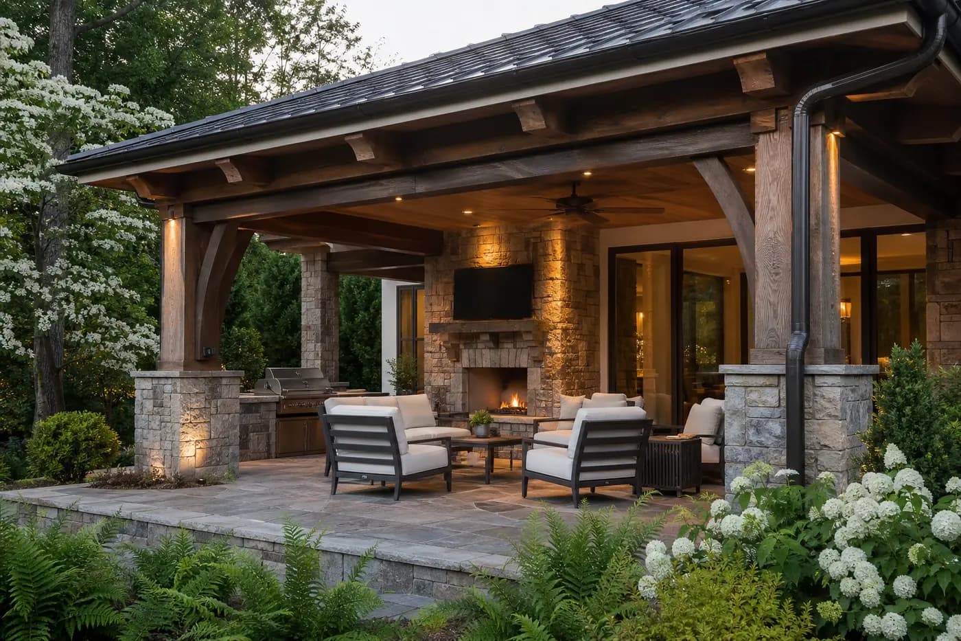 Covered stone outdoor living room with fireplace, kitchen, fan, seating, and layered landscaping